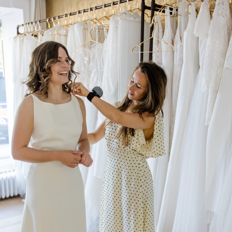 Brautkleidanprobe in der Classique Brautboutique Eine Braut strahlt und lächelt bei der Brautkleidanprobe. Inhaberin steckt das Brautkleid ab.