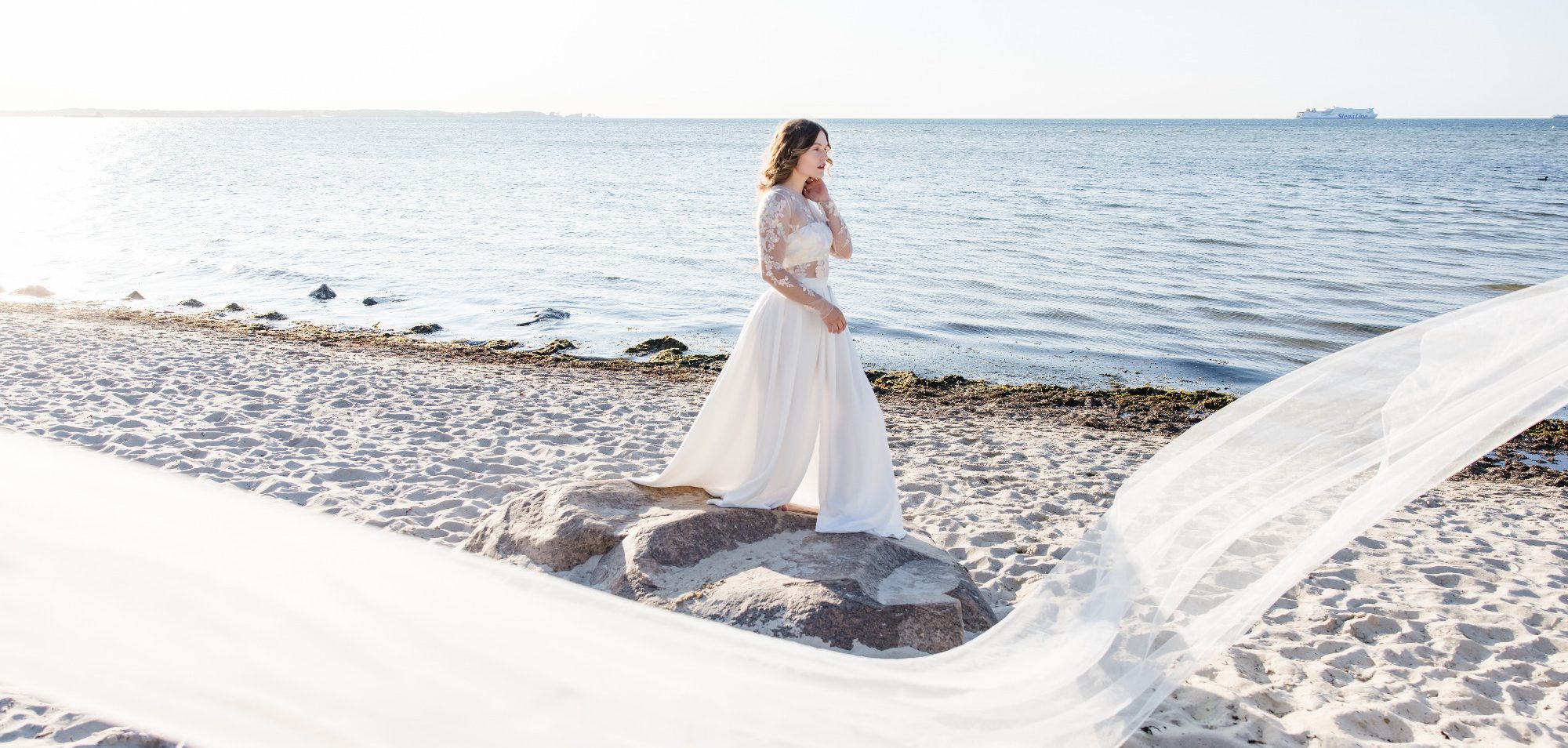 Braut am Strand in der Sonne. Die perfekte Strandhochzeit. Braut in einem Zweiteiler als Brautoutfit. Ein Crepe Rock mit Beinschlitz weht im Wind am Strand. Ein transparentes Blumenoberteil strahlt.