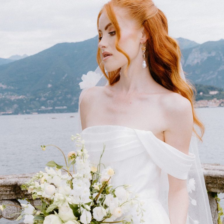 Fotografin Karina Behrendt zeigt unser trägerloses Brautkleid am Lake Como in Italien. Braut vor dem Lake Como in Italien mit roten Haaren wird seitlich in ihrem Brautkleid gezeigt. Sie hält einen Blumenstrauß.