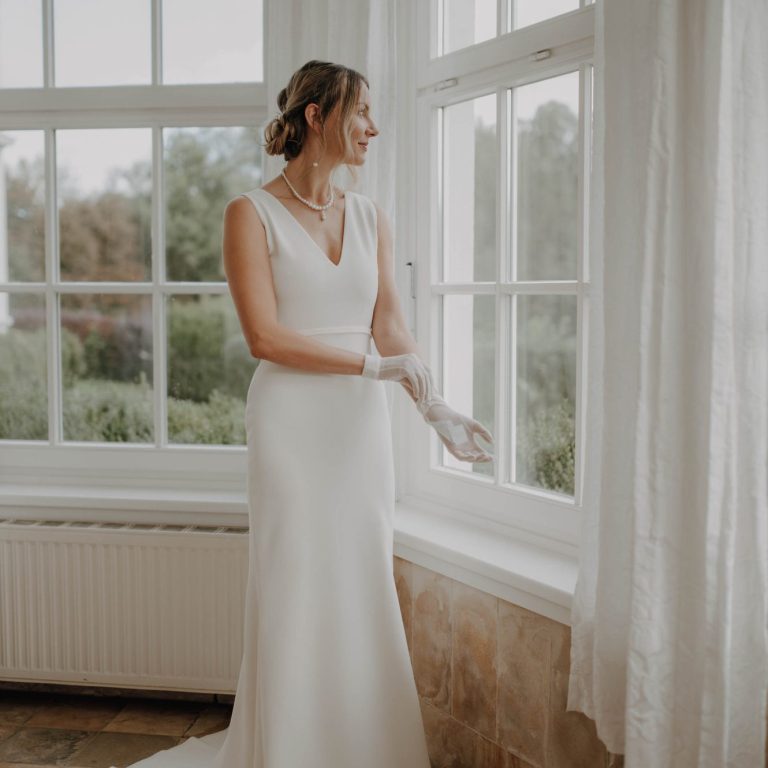 Brautkleid mit V-Ausschitt aus cleanem Stretch-Crepe. Fit&Flaire Brautkleid. Braut schaut aus dem Fenster auf die Hochzeit im Garten. Zarte Handschuhe und ein Clean Bridal Look.