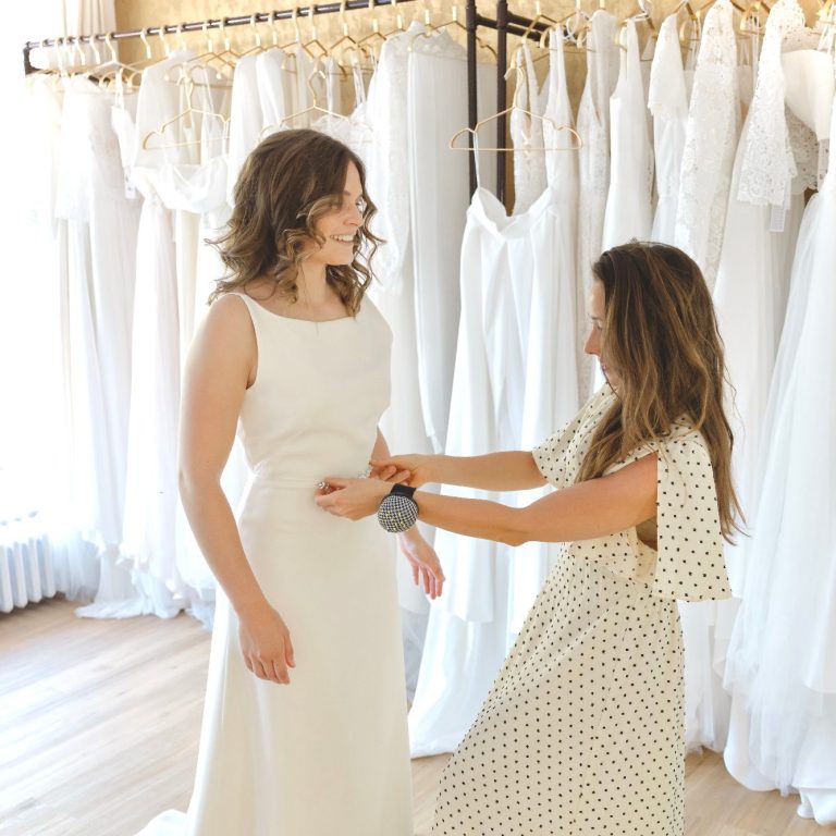 Brautkleidanprobe in Kiel Eine Braut strahlt und lächelt bei der Brautkleidanprobe. Inhaberin steckt das Brautkleid ab.