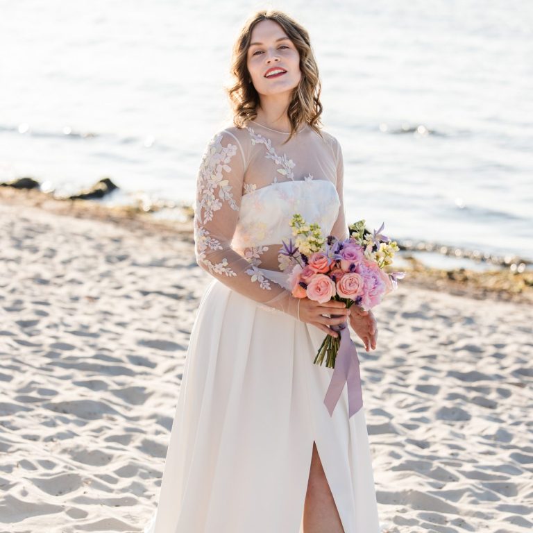 Dein Brautoutfit für deine einzigartige Strandhochzeit. Braut am Ostseestrand bei einer Strandhochzeit. Ein Oberteil mit pastellfarbigen Blumen und einen Brautstrauß in der Hand.