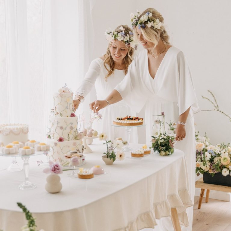 Jumper statt Brautkleid - Perfekt für die Gartenparty. Brautmode mit Jumpern. Bräute stehen vor der Candy-Bar mit Hochzeitstorte. Beide tragen Blumenkränze als Brautaccessoire.